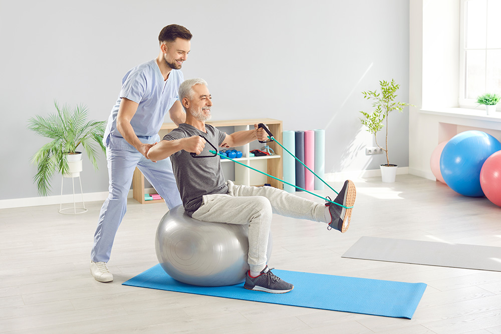 A smiling older man sits on an exercise ball, using a resistance band for a leg and arm workout, while a male physical therapist assists him in a bright, modern gym with exercise equipment in the background.