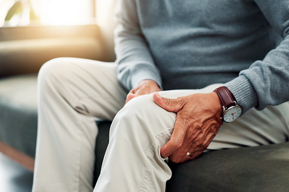 An older adult wearing a blue sweater and beige pants sits on a couch, holding their knee with one hand, suggesting discomfort or pain in the knee.