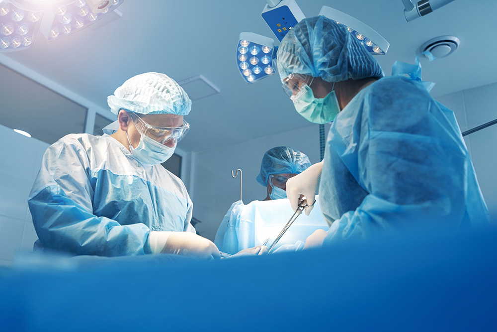 Three surgeons wearing masks, gowns, and gloves perform surgery under bright operating room lights, focusing on the procedure with surgical tools in hand.