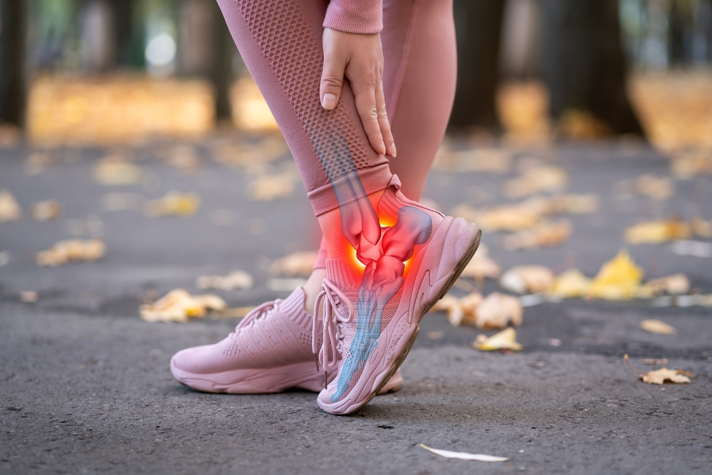 A person in pink athletic wear clutches their ankle in pain, with a glowing red and blue overlay highlighting the ankle area, indicating an injury, on a park path covered with fallen leaves.