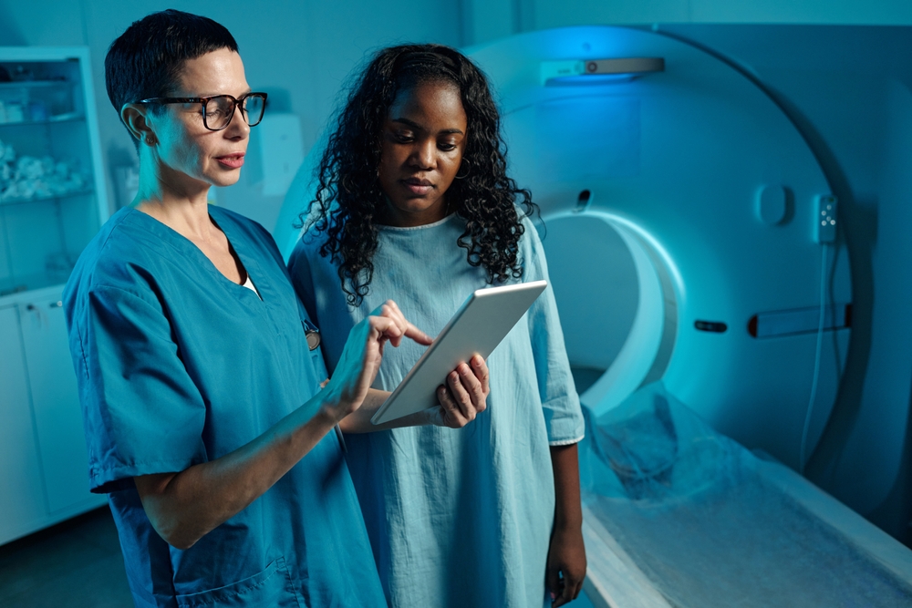 A medical professional in blue scrubs shows a patient information on a tablet beside an MRI machine in a hospital room, both focused on the screen.