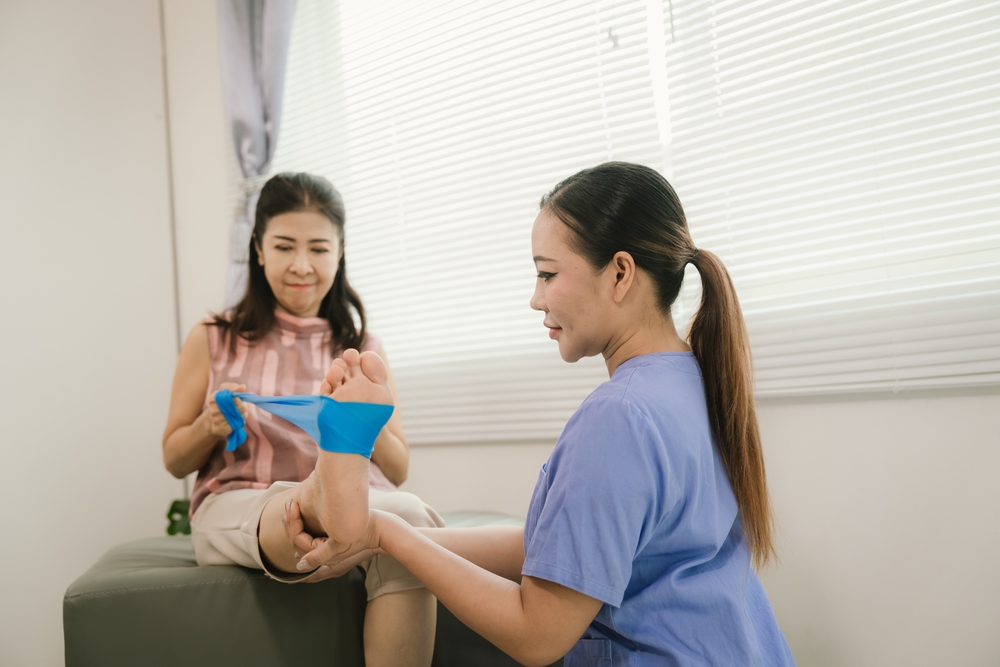 A physical therapist in blue scrubs helps a seated woman exercise her foot using a blue resistance band in a bright, clinical room with blinds.