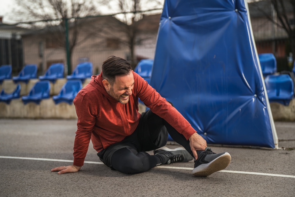 A man in athletic clothing sits on an outdoor court, holding his ankle and wincing in pain, suggesting he is injured. Blue bleacher seats and a padded pole are visible in the background.
