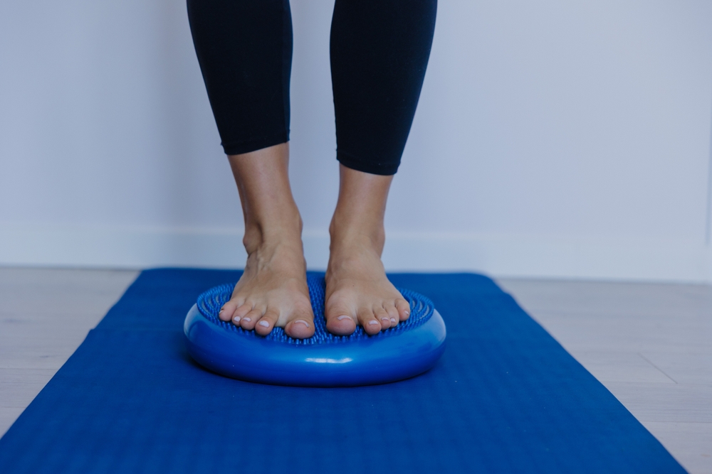 A person stands barefoot on a blue balance cushion placed on a blue exercise mat, with only their lower legs visible. They are wearing black leggings.