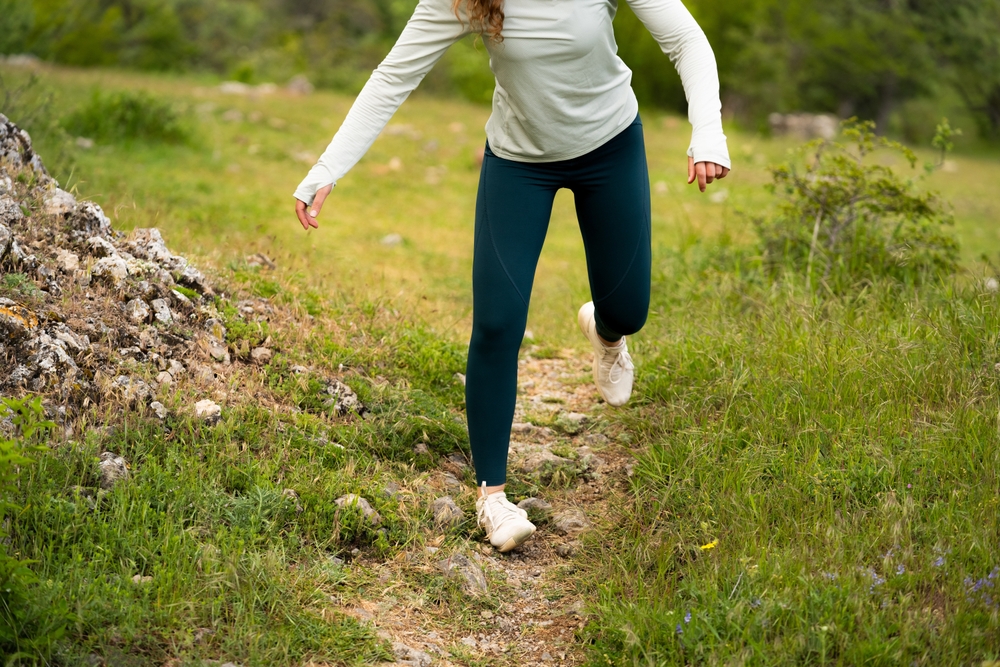 A person in a long-sleeve shirt and dark leggings is mid-step on a narrow, rocky trail in a grassy, green outdoor setting. Their head is out of frame.