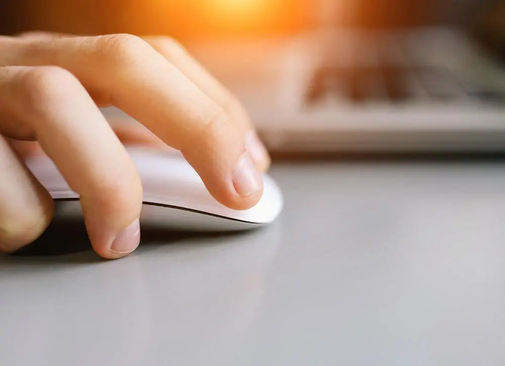 Close-up of a hand using a white computer mouse on a gray surface, with part of a laptop keyboard visible in the background and warm sunlight shining.