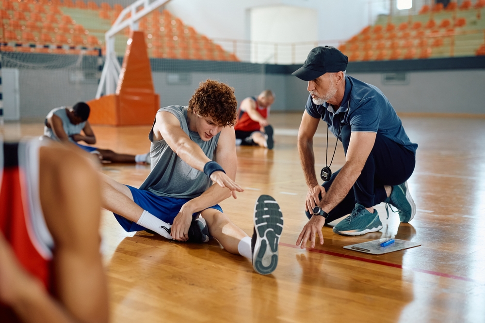 A basketball coach kneels beside a seated player who is stretching his leg on a gym floor. Other players are stretching in the background. The gym has orange seats and a basketball hoop.