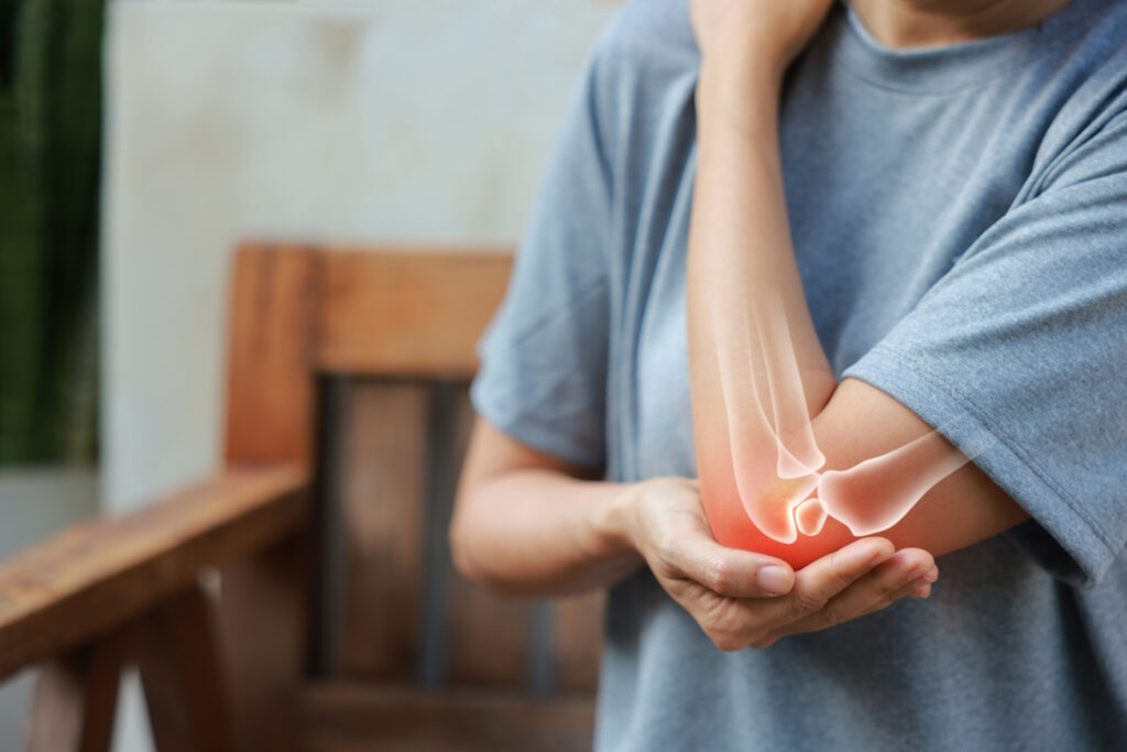 A person in a gray shirt holds their painful elbow with one hand. An illustration of elbow bones and a red glow highlight the area of discomfort, suggesting joint pain or injury.