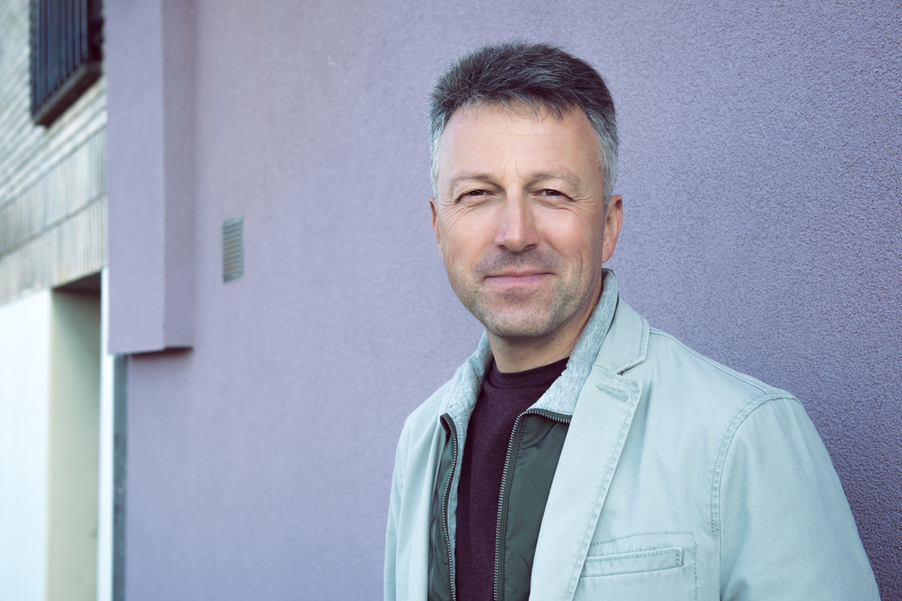 A middle-aged man with short gray hair and a slight smile stands outside against a light purple wall, wearing a light jacket over a dark shirt.