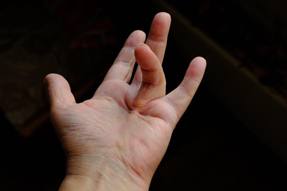 A left hand with the palm facing up; the middle finger is bent downward while the other fingers are extended, creating a gesture against a dark, blurry background.