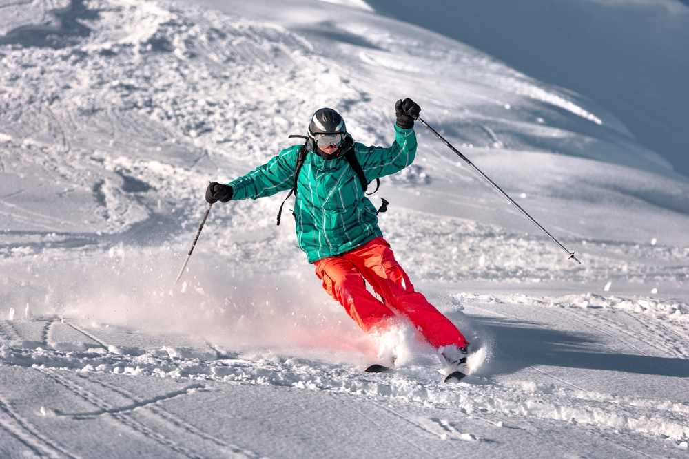 A skier in a green jacket and red pants is skiing downhill on fresh snow, wearing a helmet and goggles, with ski poles extended and snow spraying behind.