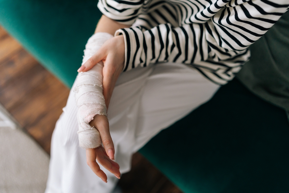 A person wearing a black and white striped shirt and white pants sits on a green couch with their hand and wrist bandaged, suggesting an injury.