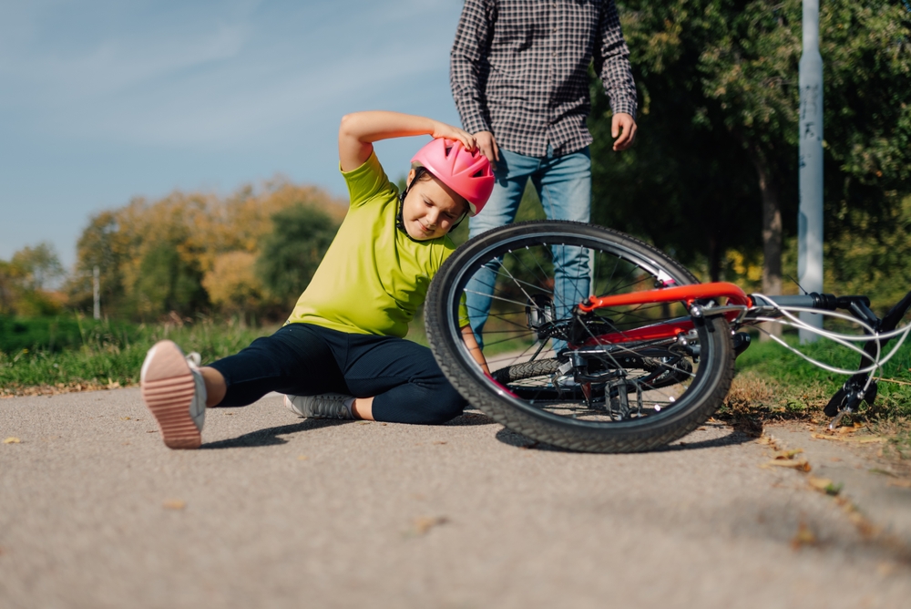 A child in a pink helmet sits on the pavement next to a fallen bicycle, holding their head in pain, while an adult stands nearby. It appears to be a sunny day in a park setting.
