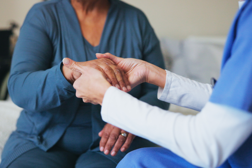 A healthcare worker gently holds the hand of an older adult, offering comfort and support. Both are seated, with attention focused on the hands, emphasizing care and compassion.