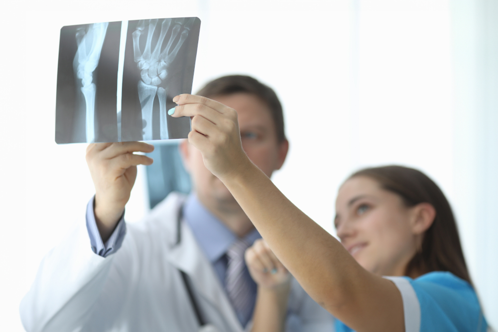 A doctor and a patient examine wrist X-ray images together, both focusing on the scans. The doctor is wearing a white coat and stethoscope, while the patient smiles and points at the X-rays.
