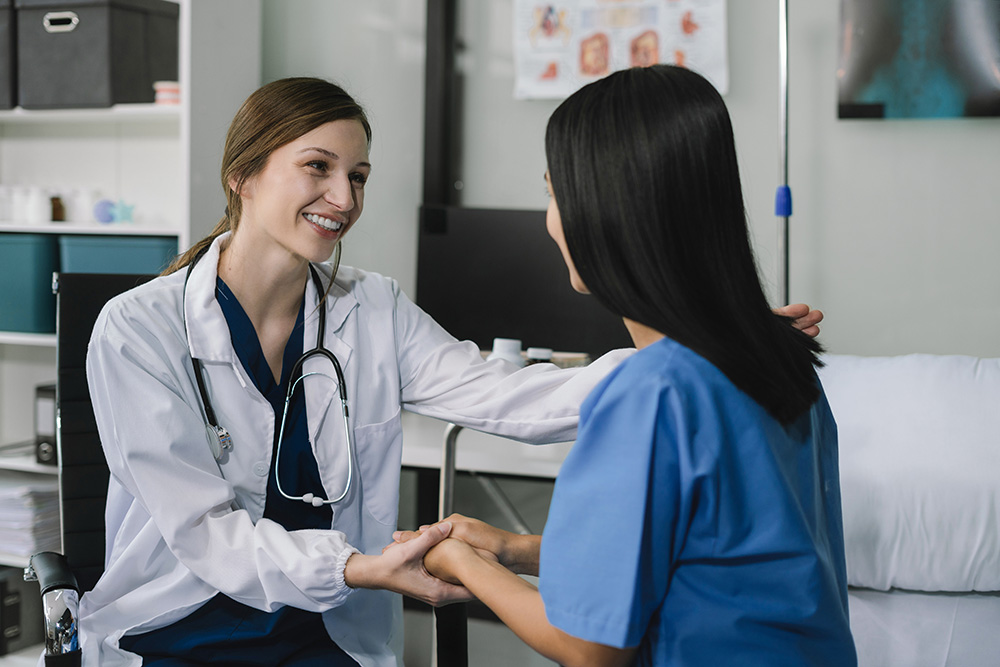 A smiling female doctor in a white coat sits and holds hands with a patient in blue scrubs, offering comfort and support in a medical office setting.
