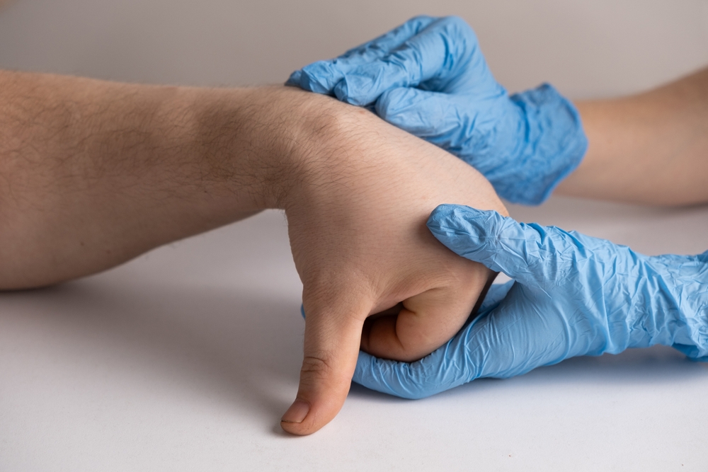 A person wearing blue medical gloves is holding and examining another persons hand, bending their thumb downward against a neutral background.