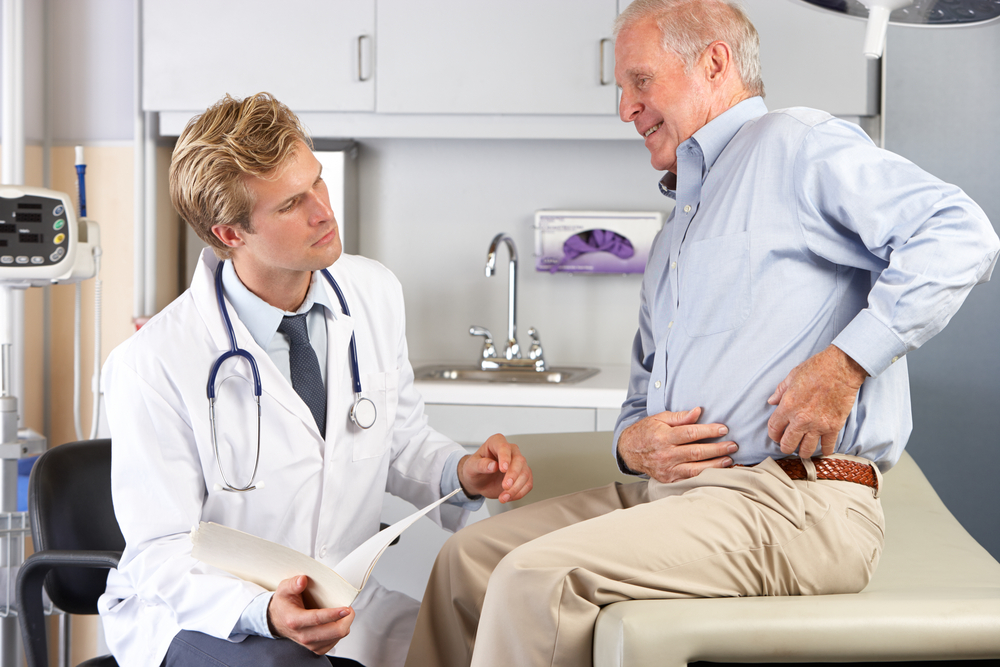 A doctor in a white coat consults with an elderly man sitting on an exam table, who appears to be experiencing hip pain and is touching his hip while talking to the doctor in a medical office.