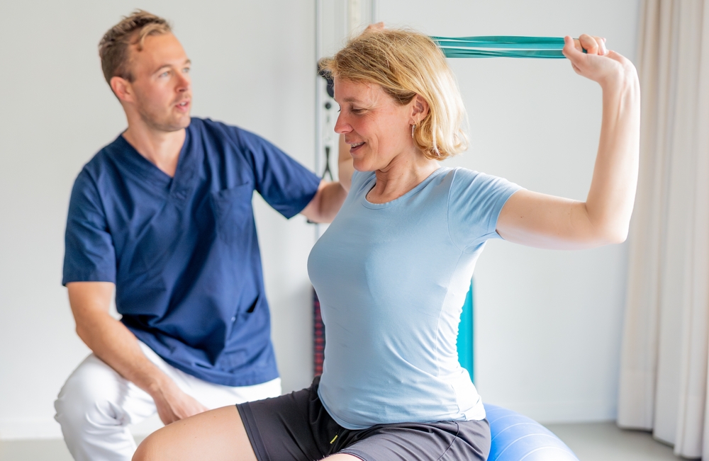 A woman sits on an exercise ball, smiling as she stretches an elastic band behind her shoulders. A physical therapist in blue scrubs guides her during the session in a bright, airy room.