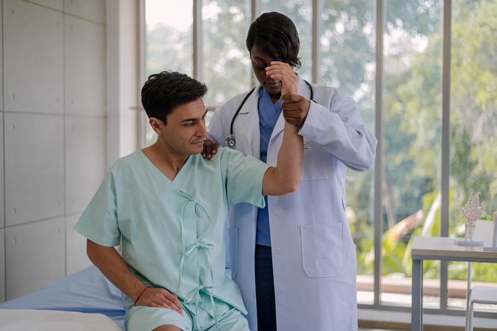 A doctor helps a patient in a hospital gown lift his arm during a physical therapy or rehabilitation session in a bright medical office with large windows.