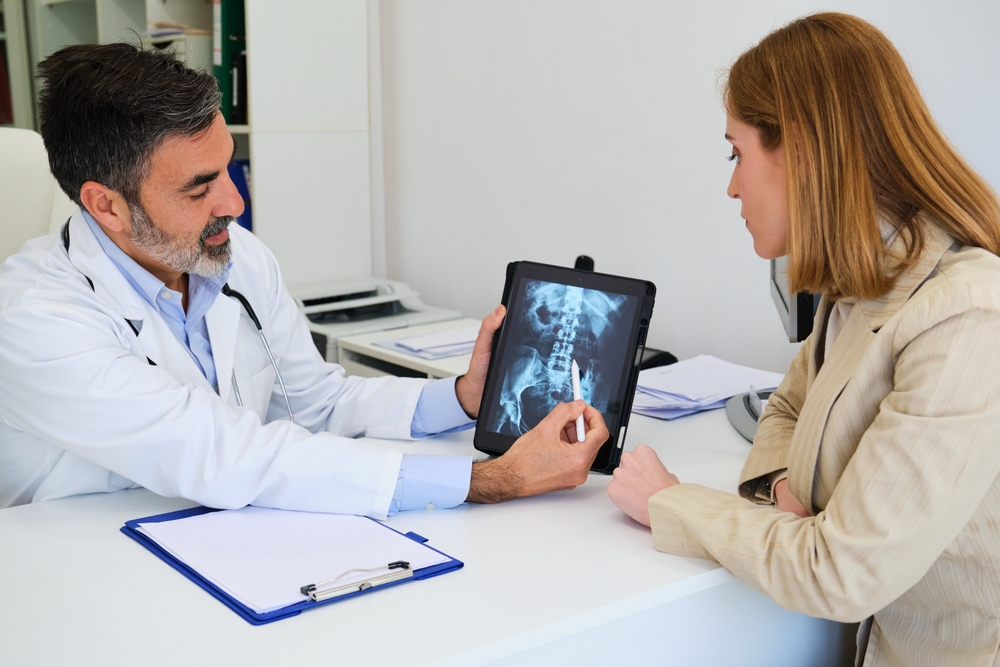 A doctor sits at a desk showing a female patient an X-ray image on a tablet, pointing to a specific area while discussing the results. Both appear focused and engaged in conversation.