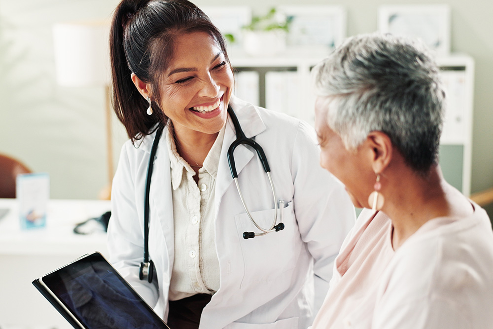 A smiling female doctor with a stethoscope talks with an older woman patient, both appearing happy and engaged during a consultation in a medical office.