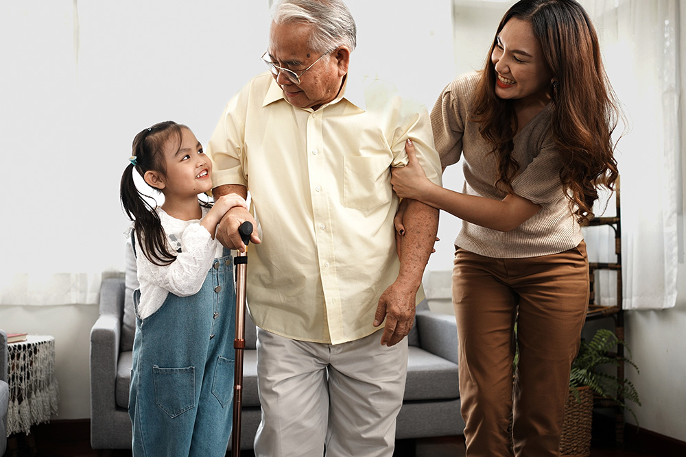 A smiling elderly man with a cane stands between a young girl and a woman, both supporting him. The girl holds his hand and smiles up at him, while the woman holds his arm and smiles warmly. They are indoors in a well-lit room.