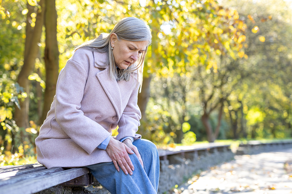 An older woman sitting on a bench outdoors, wearing a light pink coat and holding her knee with a pained expression, suggesting knee discomfort or pain. Trees with autumn foliage are in the background.