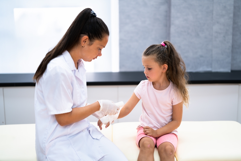 A healthcare professional wearing gloves examines the arm of a young girl sitting on an exam table. The girl looks at her arm while the professional gently checks it, both appearing focused in a medical setting.