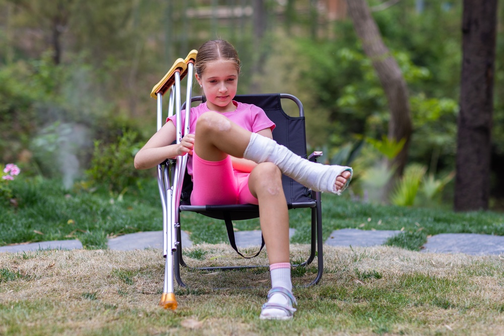A young girl with a bandaged leg sits on a folding chair outdoors, holding crutches beside her. She is wearing a pink outfit and white sandal, with a garden and trees visible in the background.