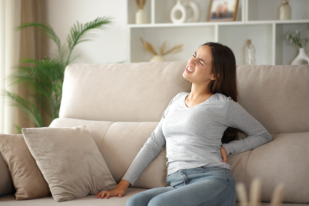 A woman sits on a beige sofa, holding her lower back with a pained expression, suggesting back pain. She is wearing a gray long-sleeve shirt and jeans. The room is decorated with neutral colors and plants.