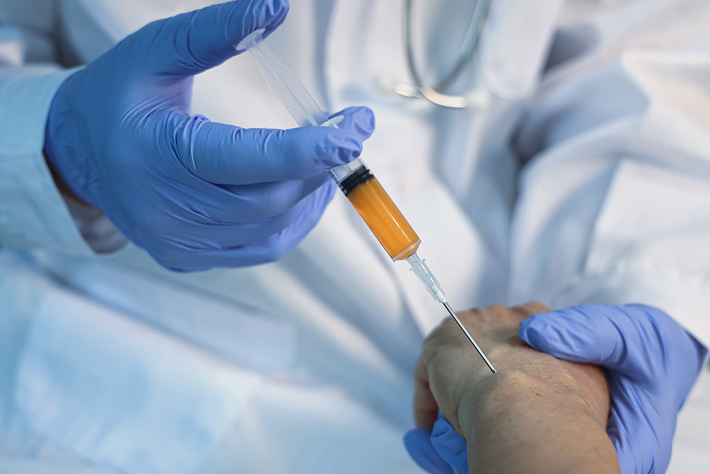 A healthcare worker wearing blue gloves injects a yellowish liquid into a patient’s hand using a syringe, with both individuals dressed in white medical attire.