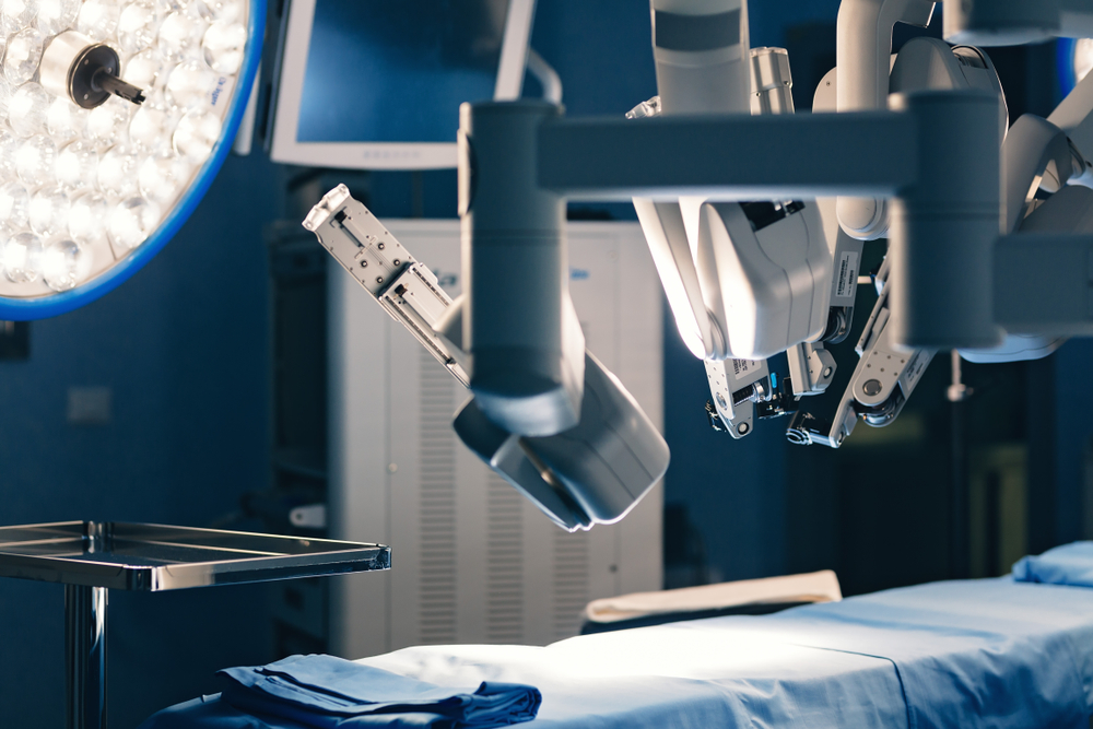 Robotic surgical arms positioned above an operating table in a modern, well-lit operating room, with medical equipment and monitors visible in the background.
