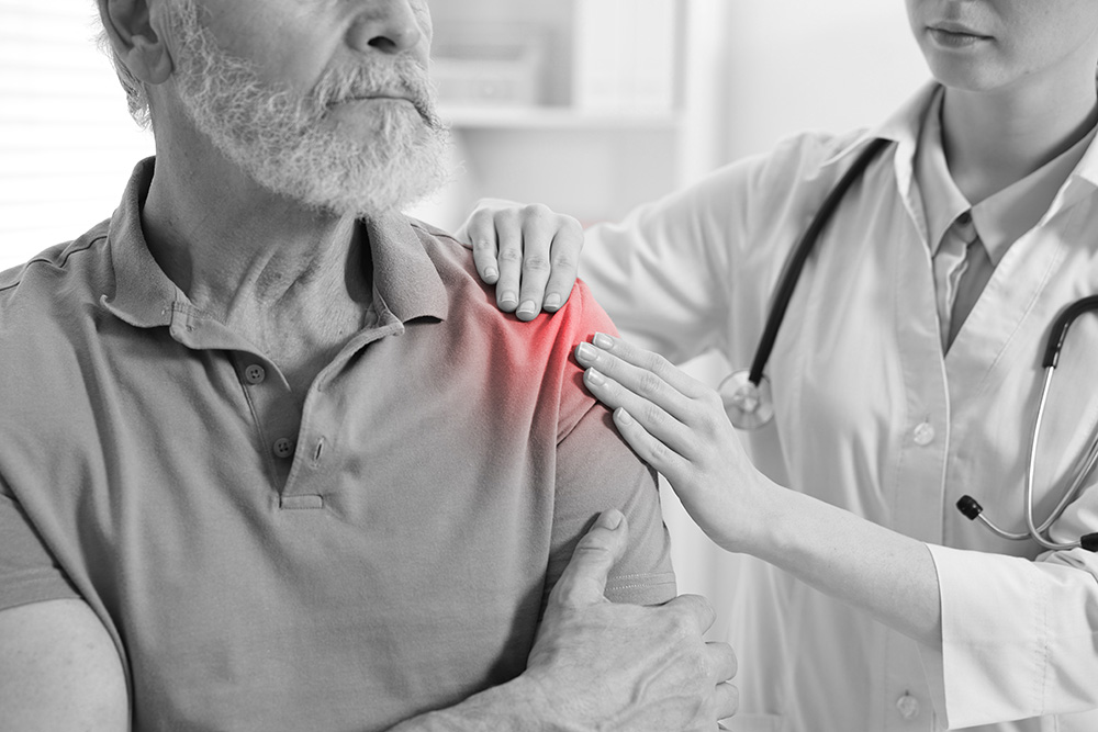 A doctor examines an older mans shoulder, which is highlighted in red to indicate pain or injury. The man looks concerned and is holding his arm, while the doctor gently touches his shoulder.