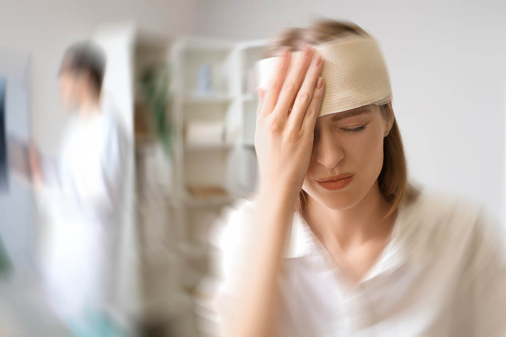 A woman with a head bandage holds her forehead in pain, eyes closed, standing in a bright medical setting with a blurred healthcare professional in the background.