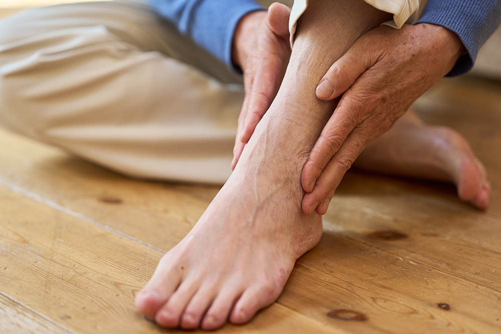 A person sitting on the floor holds their ankle with both hands, as if experiencing pain or discomfort. The focus is on the foot and ankle, with a wooden floor visible beneath them.