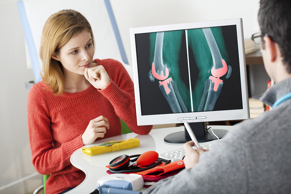 A woman in a red sweater sits across from a doctor who is showing her an X-ray of knees with artificial joints on a computer screen during a medical consultation.