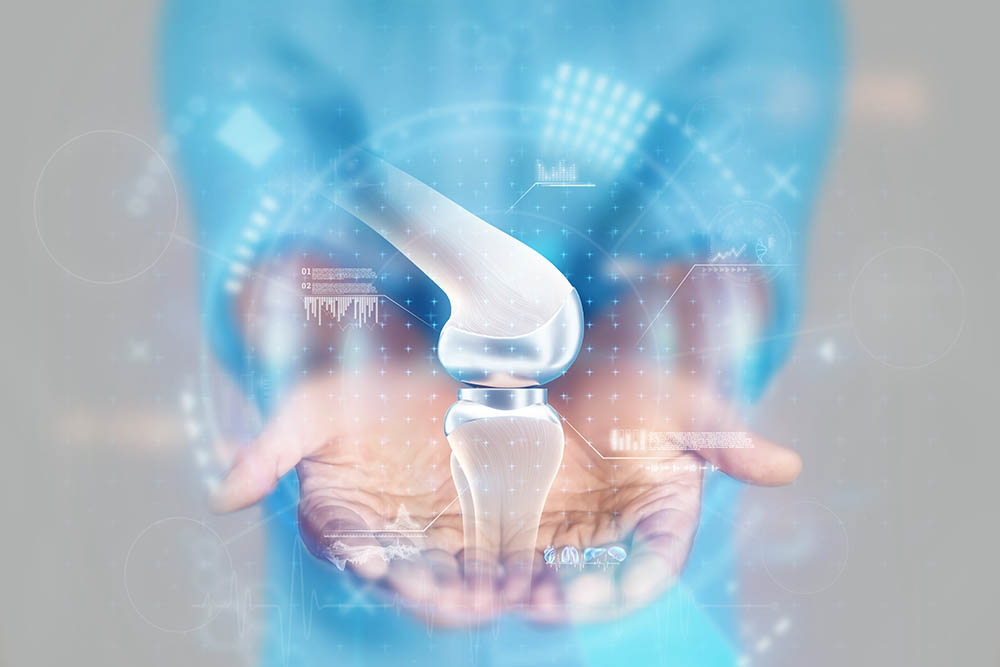 A person in blue scrubs holds out their hands, with a digital hologram of a knee joint replacement implant floating above their palms, representing advanced medical technology.