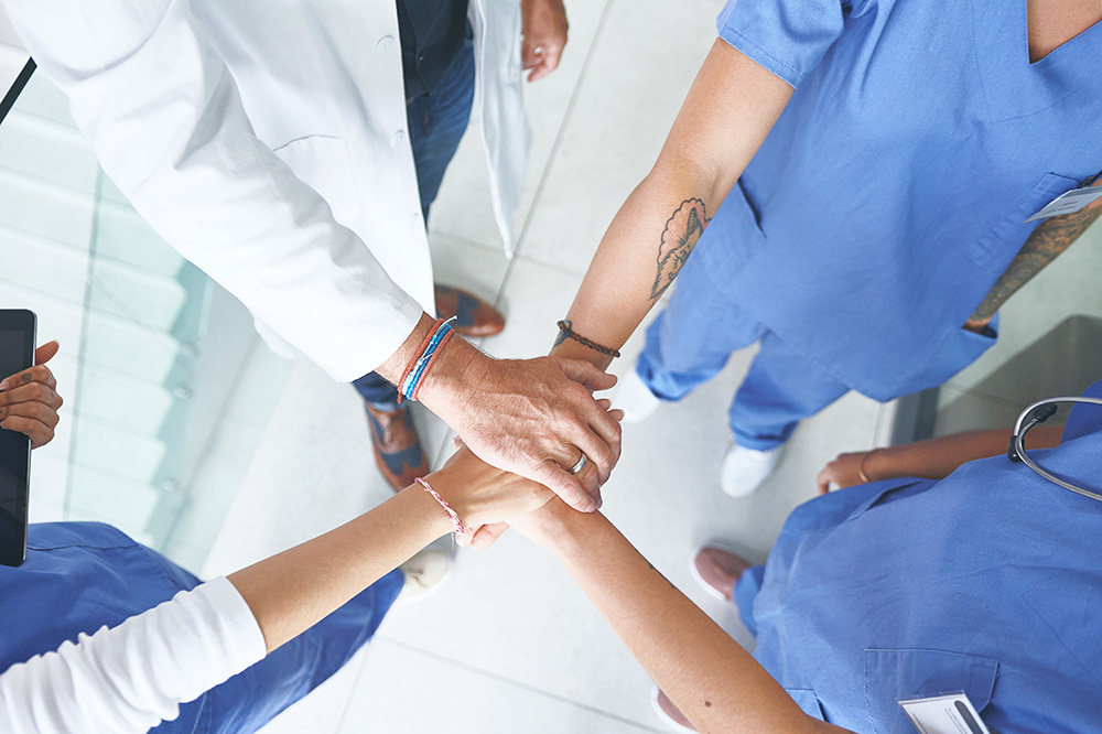 A group of healthcare professionals in scrubs and a lab coat stand in a circle, stacking their hands together in a gesture of teamwork and unity.