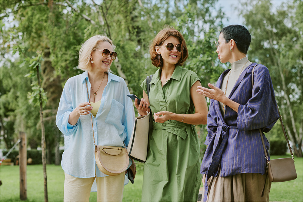 Three women wearing sunglasses and stylish outfits walk together outdoors, smiling and talking, with trees and greenery in the background.