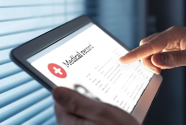 A person uses a tablet to view an electronic medical record, with one finger pointing at the screen. The display shows health information and a red medical symbol. Blue window blinds are visible in the background.