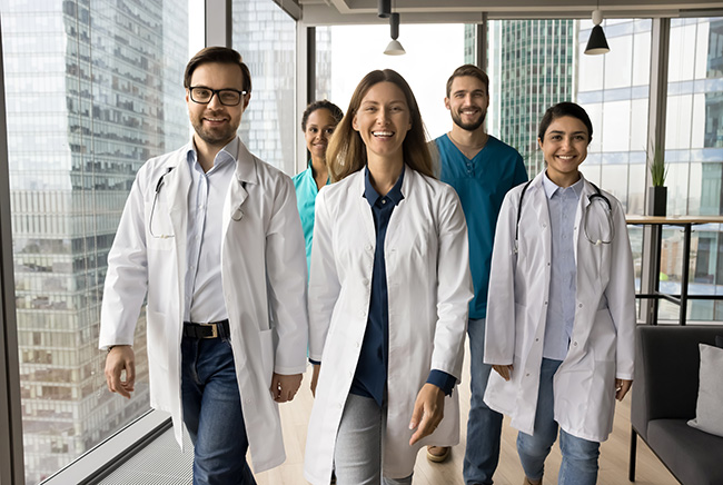 A diverse group of five smiling medical professionals, wearing lab coats and scrubs, walk confidently down a bright hallway with large windows and city buildings visible outside.