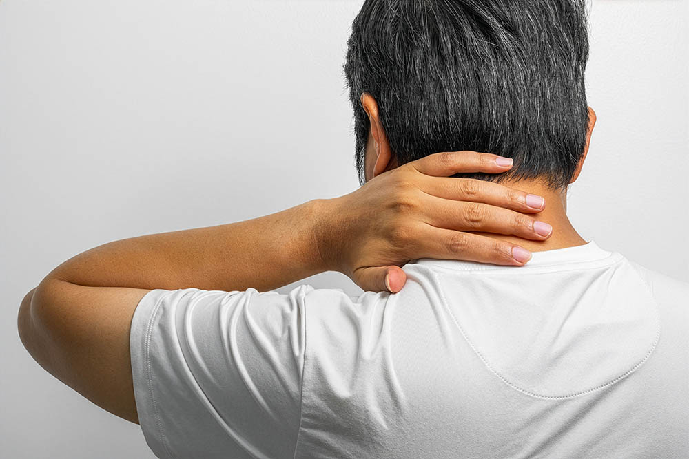 A person wearing a white shirt is shown from behind, holding the back of their neck with one hand, suggesting neck pain or discomfort.