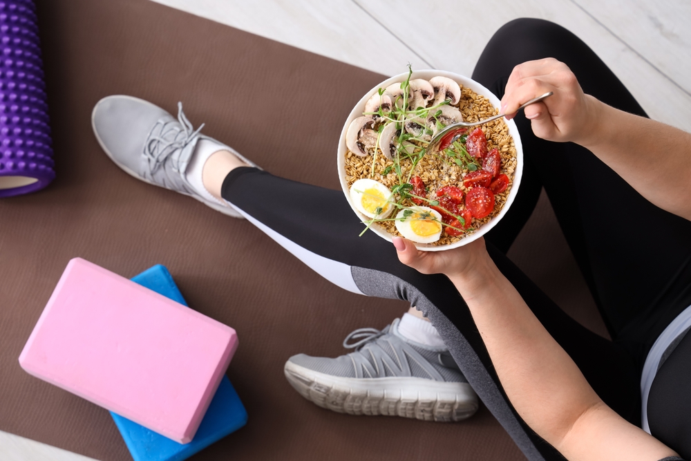 Person in workout attire sits on a yoga mat next to a foam roller and yoga blocks, holding a bowl of healthy food with eggs, tomatoes, mushrooms, and greens, eating with a fork.