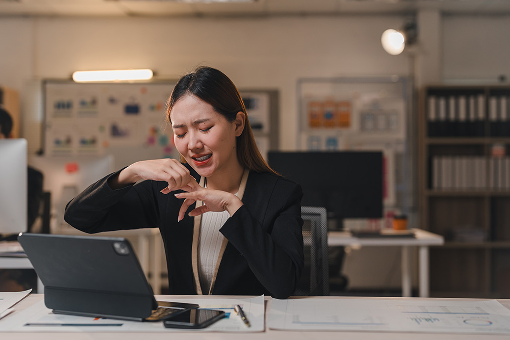 A woman in a business suit sits at a desk with a tablet, appearing frustrated or upset, holding her hand to her mouth while working in an office environment.