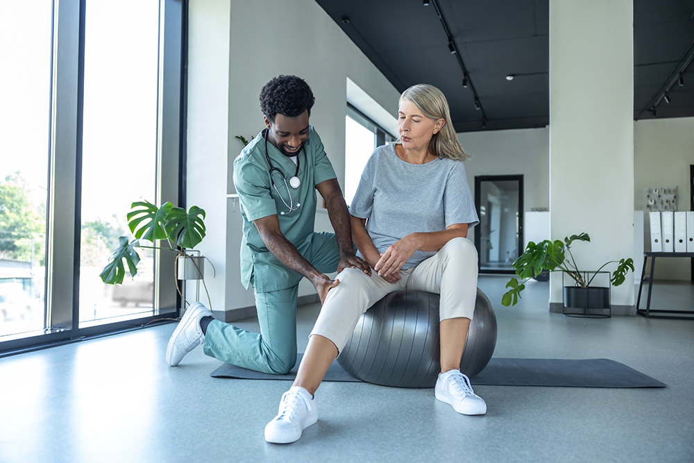 A physical therapist kneels beside a seated older woman on an exercise ball, gently examining her knee in a bright, modern rehab facility with large windows and potted plants.