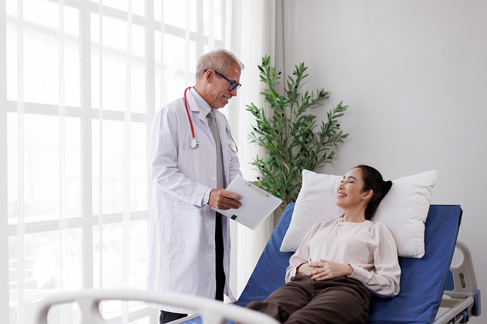 A doctor wearing a white coat and stethoscope talks to a smiling patient lying on a hospital bed in a bright, modern room with a large window and a potted plant.
