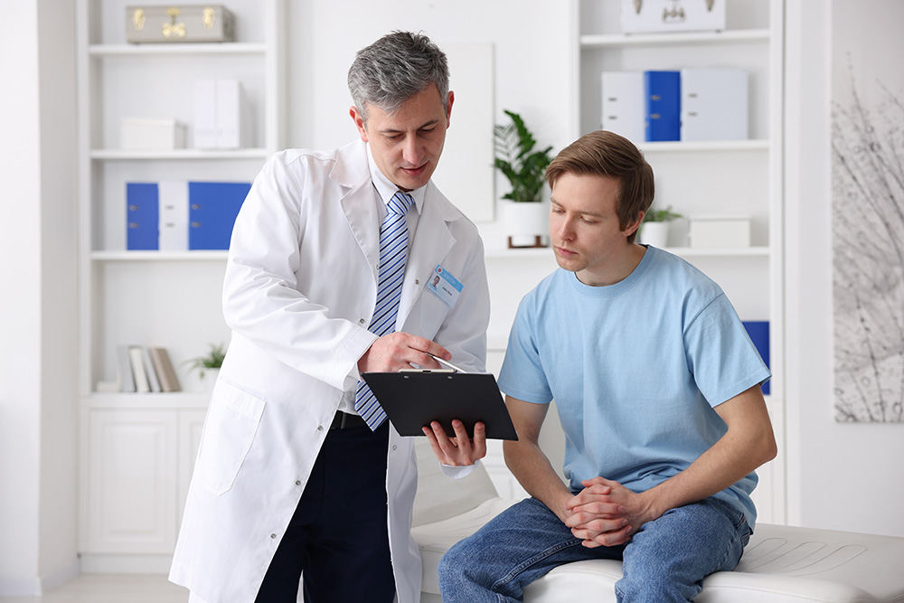 A doctor in a white coat explains notes on a clipboard to a seated male patient in a light blue shirt, inside a bright, modern medical office.