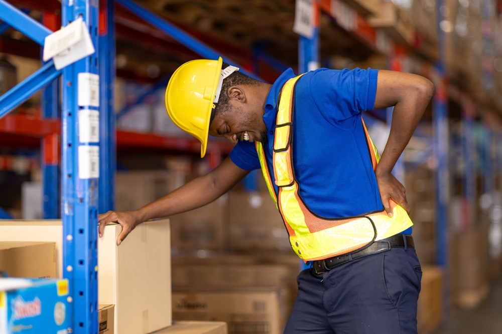 A warehouse worker wearing a yellow hard hat and safety vest stands by a shelf, holding his lower back in pain, suggesting he may have injured himself while lifting boxes.