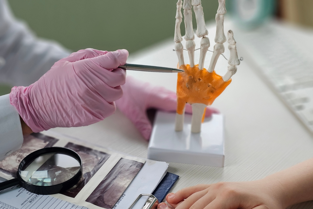 A person wearing pink gloves points at a model of a human hand skeleton, highlighting the bones, while another persons hand rests nearby on a desk with medical documents and x-rays.